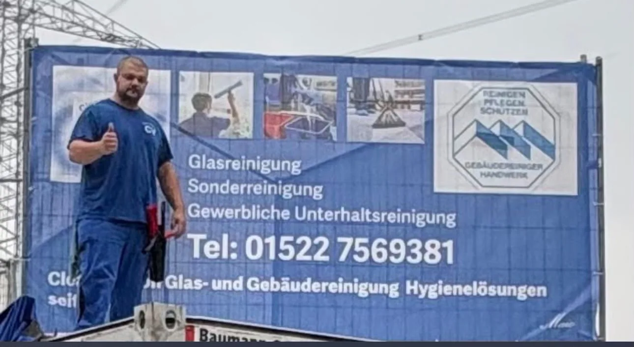 Mitarbeiter von CleanVersion mit erhobenem Daumen vor einem blauen Bauzaun-Banner mit Glasreinigung, Sonderreinigung, gewerblicher Unterhaltsreinigung und Telefonnummer